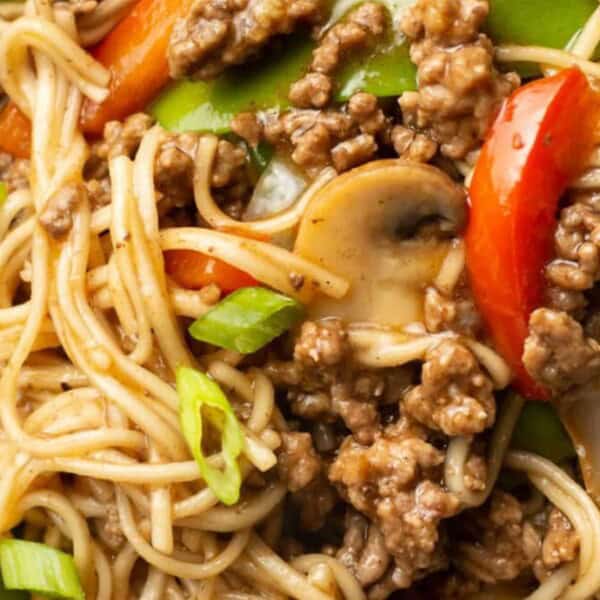 A close-up of a bowl of stir-fried noodles with ground beef, red and green bell peppers, mushrooms, and sliced green onions.