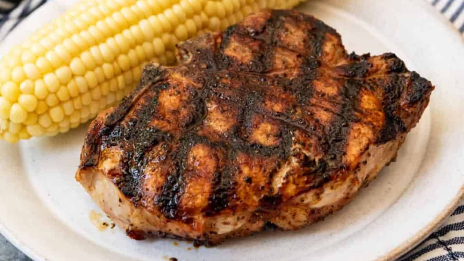 Grilled pork chop with a charred grill pattern, served alongside a cob of sweet corn on a plate.