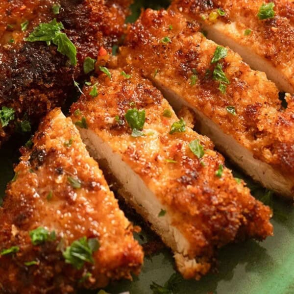 Sliced breaded and fried chicken garnished with chopped parsley on a green plate, with a lemon wedge in the corner.