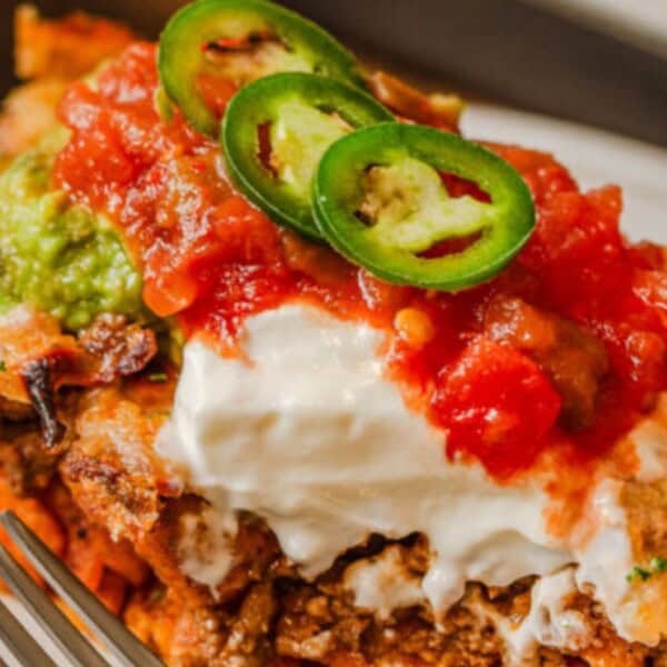 A savory dish featuring layered meat, topped with sour cream, salsa, guacamole, and sliced jalapeños on a white plate.