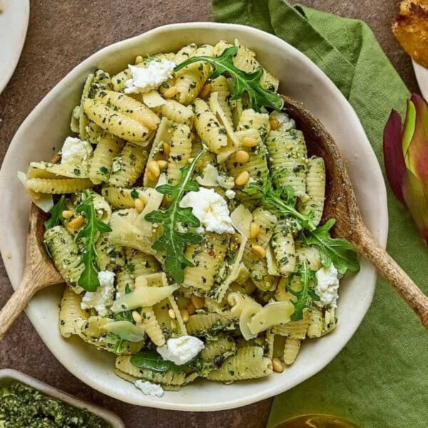A bowl of pasta with green pesto, arugula, ricotta, and pine nuts, surrounded by plates of pasta, bread, pine nuts, and pesto.