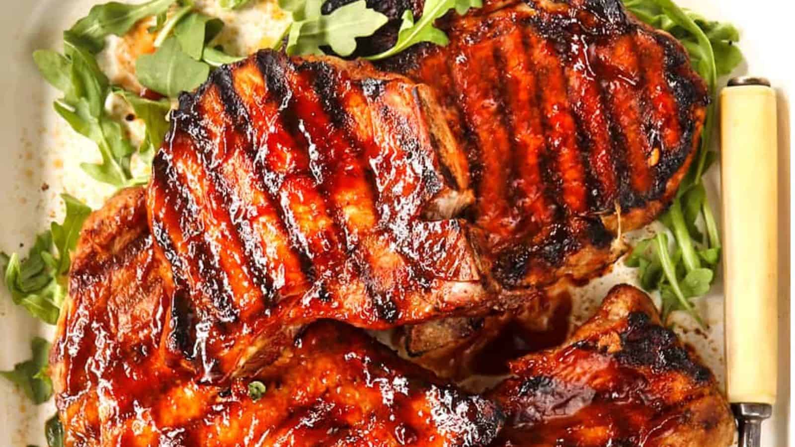 Grilled, glistening pork chops with caramelized glaze, resting on a bed of fresh arugula, served with a rustic knife.