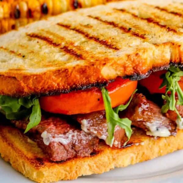 Grilled sandwich filled with steak, fresh tomatoes, and arugula on toasted bread, accompanied by a piece of charred corn.