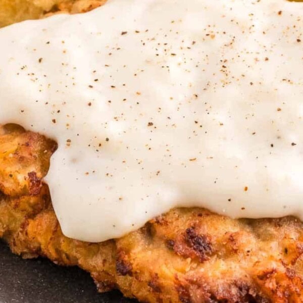 A close-up of crispy fried chicken covered in creamy white gravy, served on a dark plate with a side of green beans.