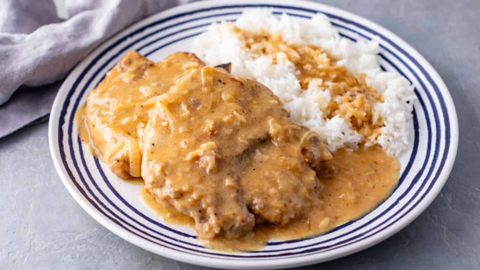 A plate of tender pork chops smothered in savory gravy, served with fluffy white rice and garnished with a striped blue rim.