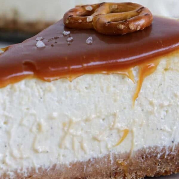 A slice of cheesecake with a caramel drizzle and pretzel pieces sits on a plate. The cheesecake has a golden graham cracker crust.