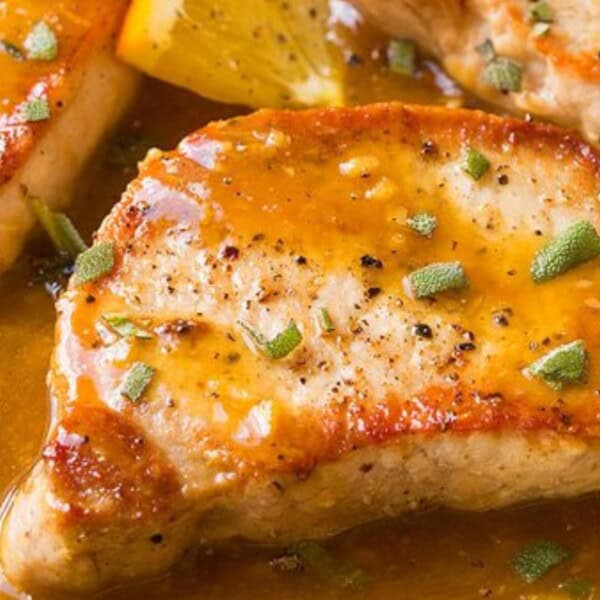 Pan-seared pork chops topped with sauce and chopped herbs, served next to fresh orange wedges.