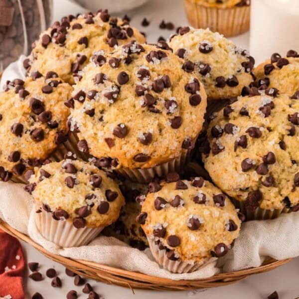 A wicker basket filled with chocolate chip muffins, sprinkled with salt, surrounded by chocolate chips and milk in glass bottles.