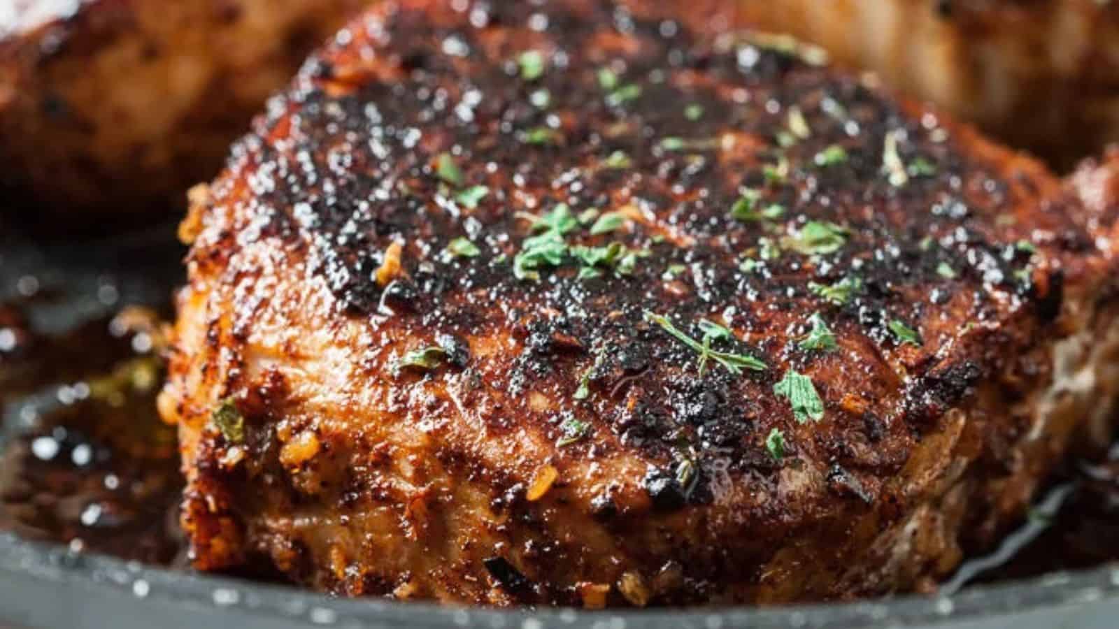 A close-up of a seared, juicy pork chop, garnished with herbs, showcasing a crispy blackened crust and glistening juices in a skillet.