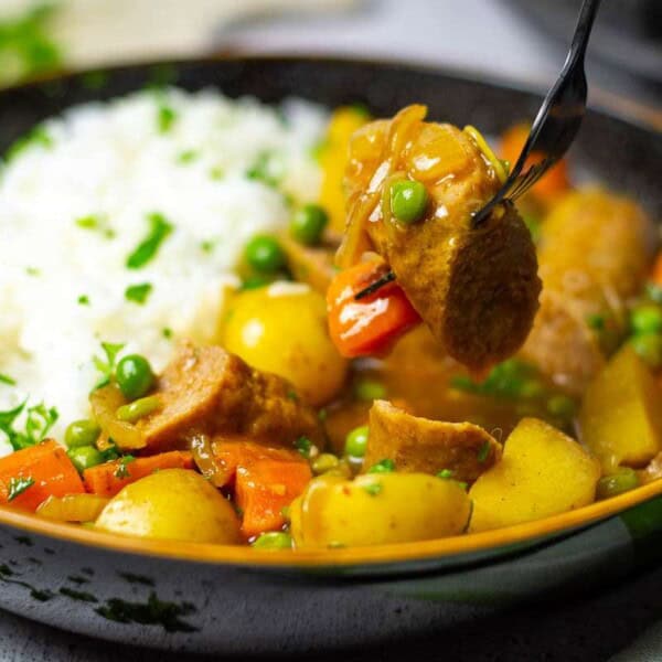 A bowl of hearty stew with chunks of meat, potatoes, carrots, and peas, served with fluffy white rice and garnished with herbs.