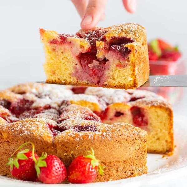 A slice of strawberry cake is being lifted with a knife from a whole cake on a white plate, with fresh strawberries beside it.