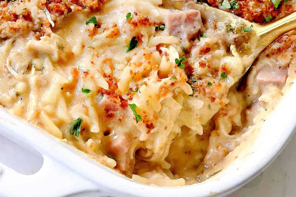 A close-up of a creamy baked casserole topped with melted cheese, small pieces of ham, and fresh herbs, served in a white baking dish with a golden spoon scooping out a portion.