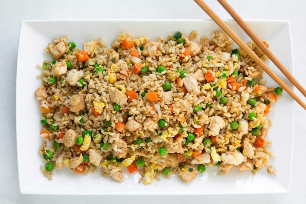 A rectangular white plate with chicken fried rice, containing diced chicken, scrambled eggs, peas, carrots, corn, and rice. Brown wooden chopsticks rest on the upper right edge of the plate.
