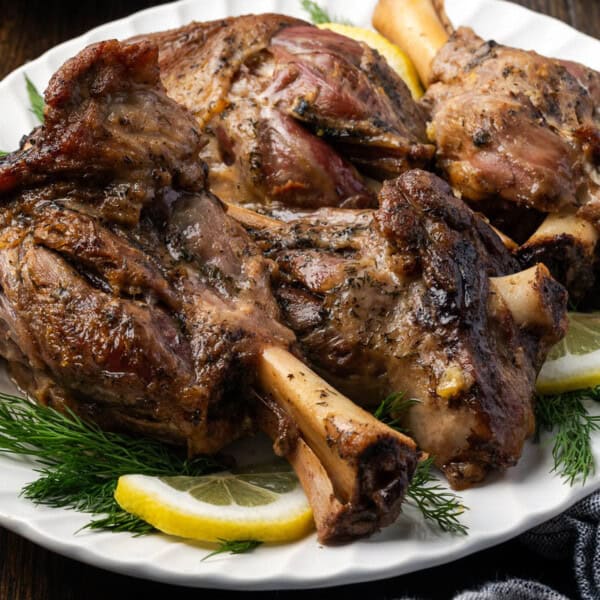 A white plate with several cooked lamb shanks, garnished with lemon slices and fresh dill, on a wooden table.