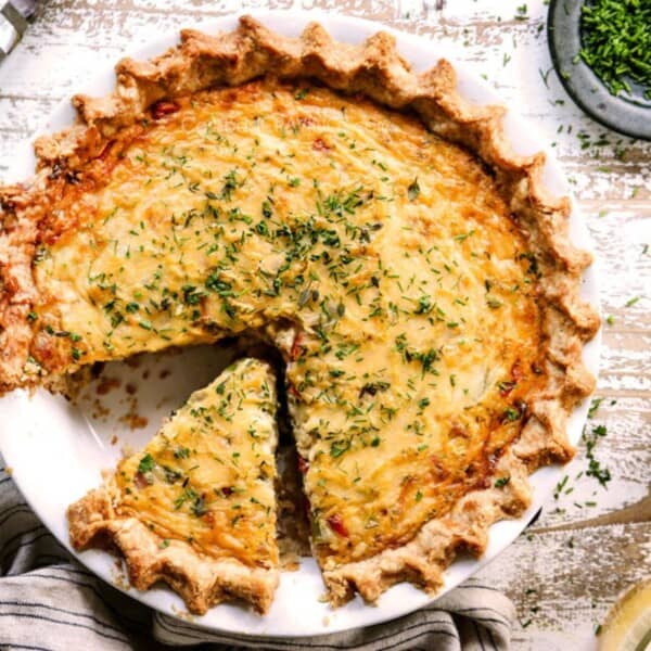 A baked quiche with a golden crust sits on a table; one slice is missing and chopped herbs are sprinkled on top.