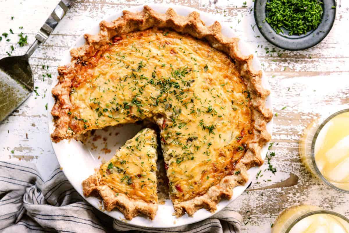 A baked quiche with a golden, flaky crust sits on a white plate. One slice is being served. The quiche is topped with chopped herbs, and a small bowl of extra herbs and two drinks are nearby on a rustic table.