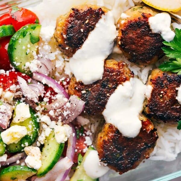 A glass container with rice, four grilled meatballs topped with white sauce, and a sprig of parsley. On the side, a salad with cucumber, cherry tomatoes, red onion, feta cheese, and herbs. A lemon wedge is placed on the rice.