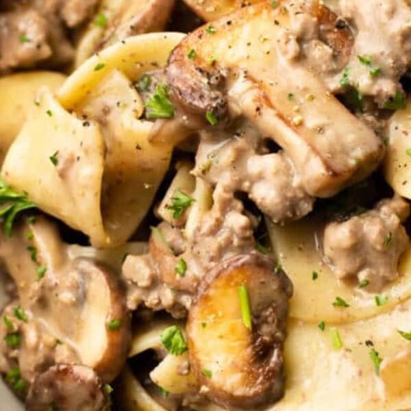 A close-up of creamy beef stroganoff featuring wide egg noodles, browned beef, sautéed mushrooms, and garnish of fresh parsley. The rich, creamy sauce coats the ingredients, visible within a white bowl.