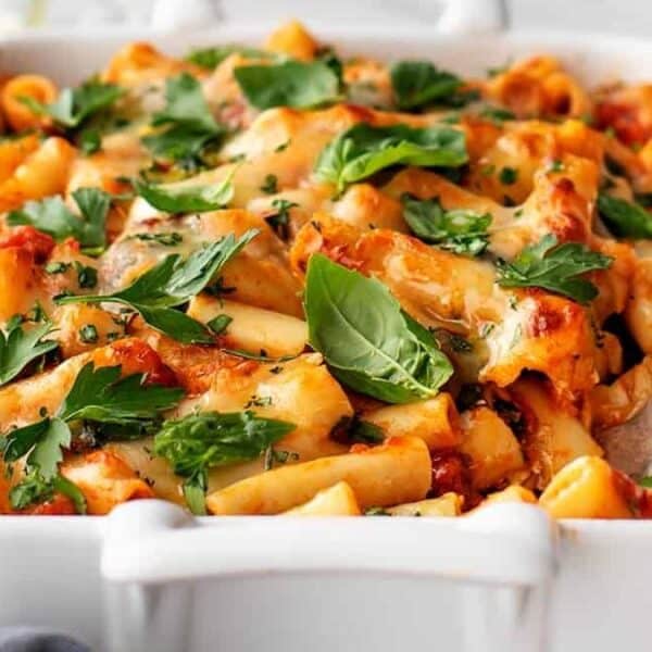 Baked ziti topped with melted cheese and fresh basil leaves in a white casserole dish, with a serving spoon on the side.