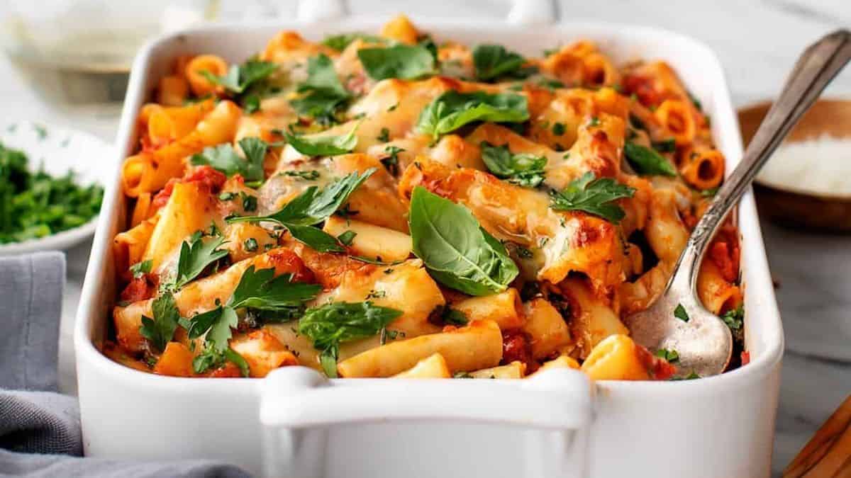 Baked ziti topped with melted cheese and fresh basil leaves in a white casserole dish, with a serving spoon on the side.