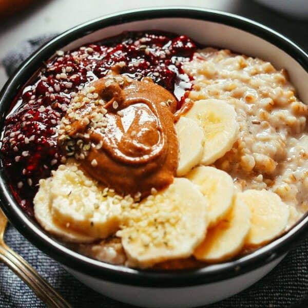 A bowl containing oatmeal, banana slices, berry jam, peanut butter, and sprinkled seeds sits beside a wooden spoon on a dark cloth.