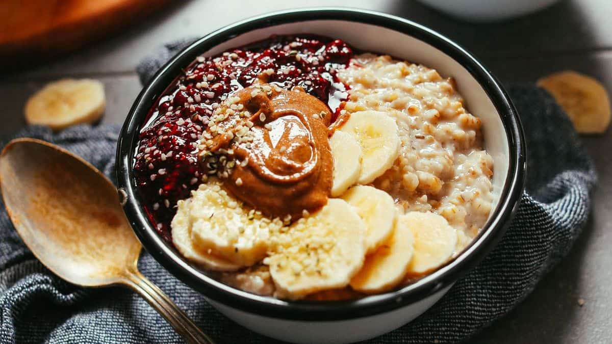 A bowl containing oatmeal, banana slices, berry jam, peanut butter, and sprinkled seeds sits beside a wooden spoon on a dark cloth.