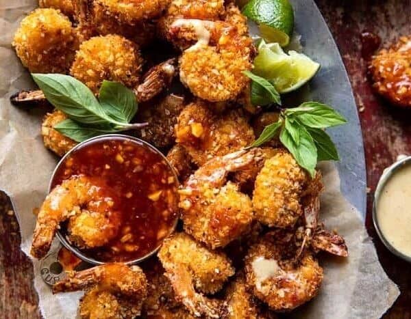 Breaded fried shrimp arranged on parchment paper, garnished with fresh basil leaves and lime wedges, accompanied by a small bowl of red dipping sauce.