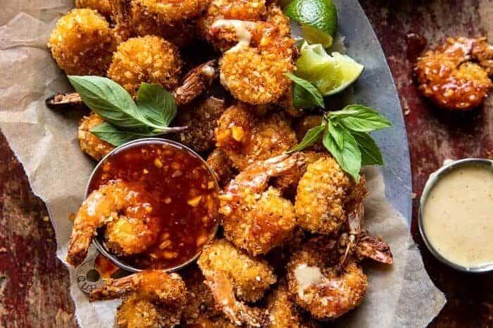 Breaded fried shrimp arranged on parchment paper, garnished with fresh basil leaves and lime wedges, accompanied by a small bowl of red dipping sauce.