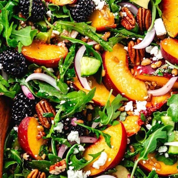 A white bowl filled with arugula, peach slices, blackberries, avocado pieces, red onion rings, pecans, and crumbled cheese. Wooden salad servers rest in the bowl.