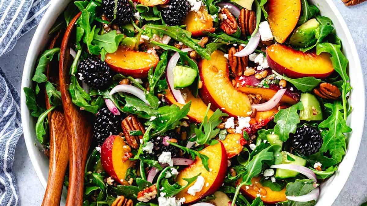 A white bowl filled with arugula, peach slices, blackberries, avocado pieces, red onion rings, pecans, and crumbled cheese. Wooden salad servers rest in the bowl.