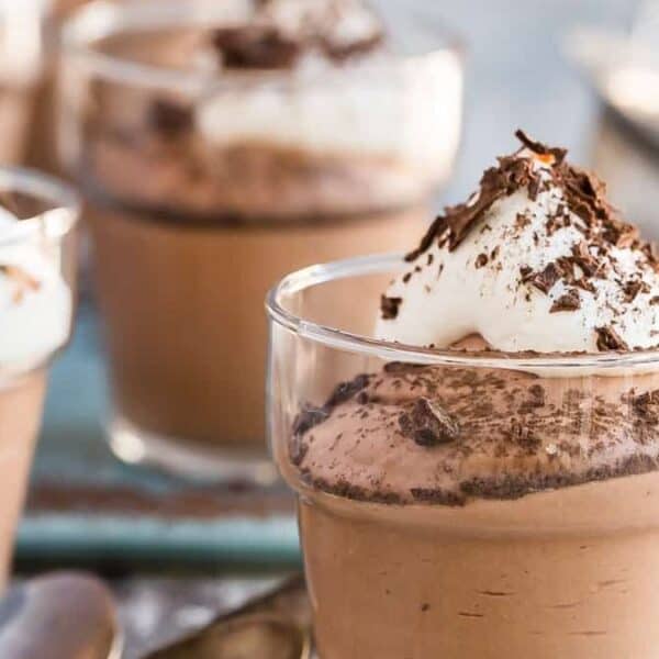 Four glass cups filled with chocolate mousse, each topped with whipped cream and chocolate shavings, are arranged on a table with spoons.