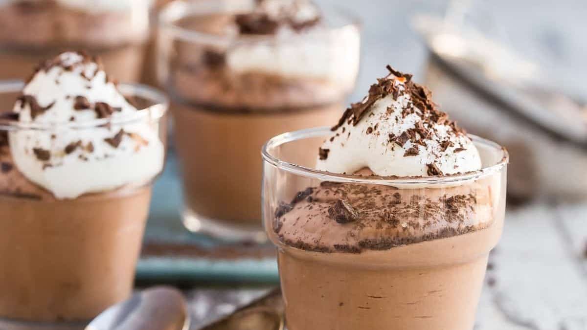 Four glass cups filled with chocolate mousse, each topped with whipped cream and chocolate shavings, are arranged on a table with spoons.