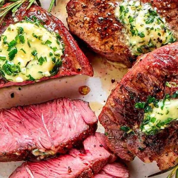 Three filet mignon steaks topped with herb butter and rosemary, arranged on a white plate; one steak is sliced to show its pink interior.