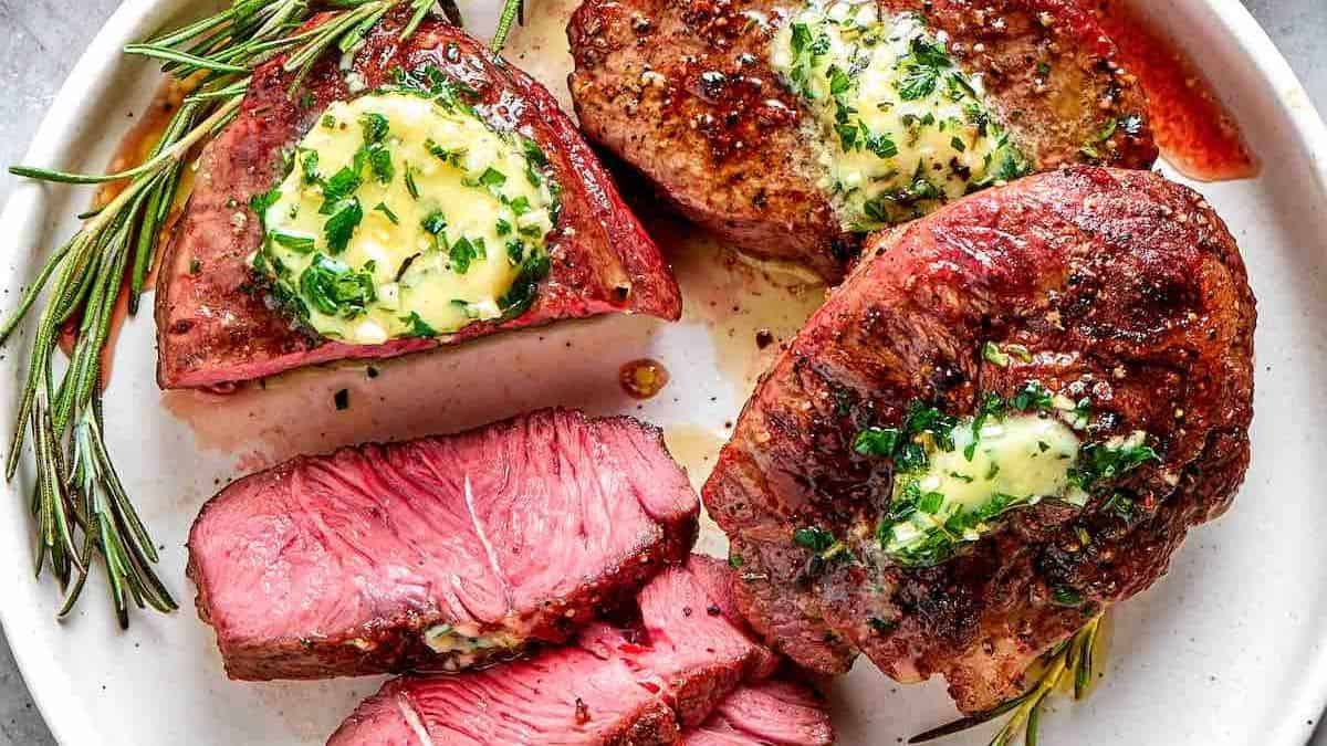 Three filet mignon steaks topped with herb butter and rosemary, arranged on a white plate; one steak is sliced to show its pink interior.