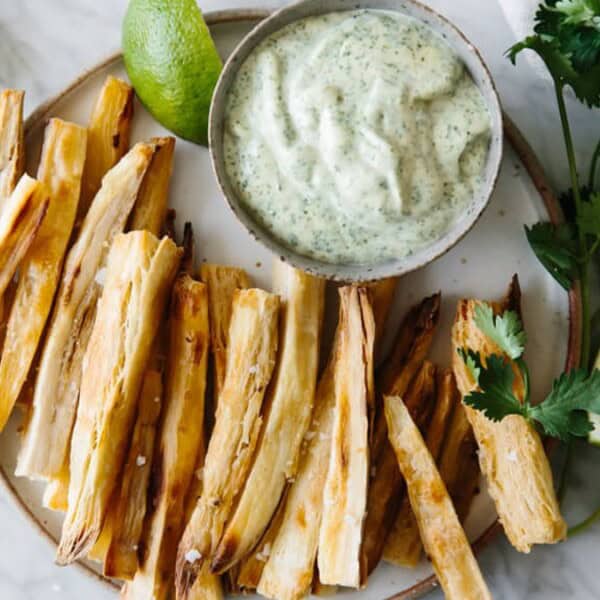 Plate of crispy yuca fries with a creamy green dipping sauce. A few lime wedges and fresh cilantro are on the side. Everything is set on a marble surface.