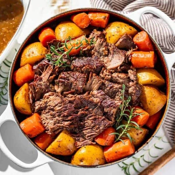 Chunks of beef, potatoes, and carrots garnished with rosemary are served in a white Dutch oven, placed next to a bowl of gravy and a wooden spoon.
