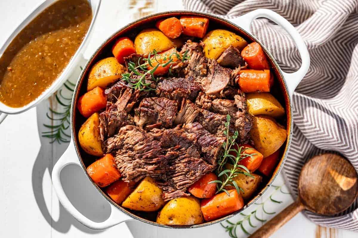 Chunks of beef, potatoes, and carrots garnished with rosemary are served in a white Dutch oven, placed next to a bowl of gravy and a wooden spoon.