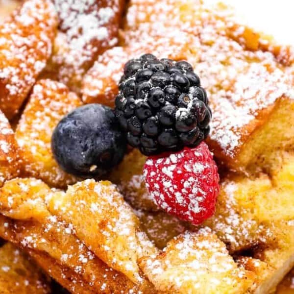 A close-up of a serving of bread pudding topped with a dusting of powdered sugar. Three berries—blackberry, blueberry, and raspberry—are placed on top for garnish. The dish has a golden, toasted appearance.