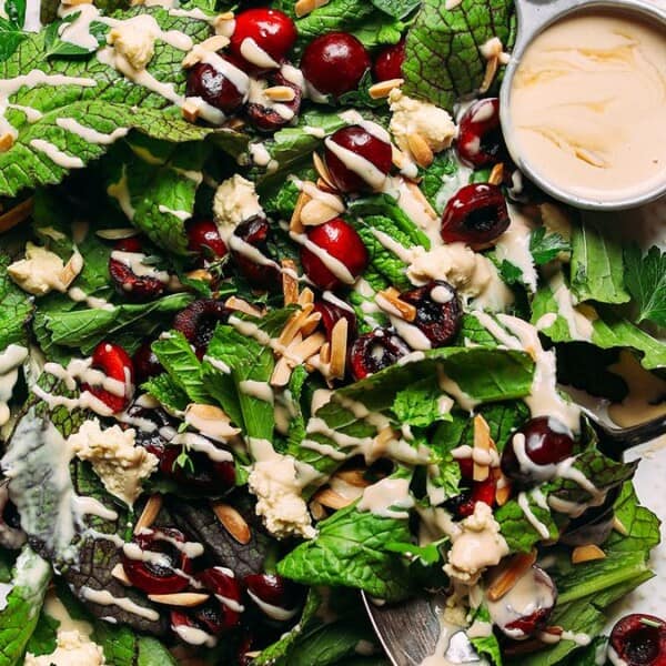 A fresh salad on a white plate featuring leafy greens, cherries, almonds, and crumbled cheese, drizzled with a creamy dressing. A hand holds a fork and spoon, about to serve. A small bowl of dressing is on the side.