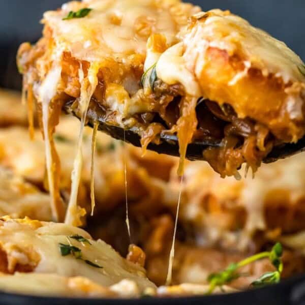 A skillet filled with slices of bread topped with melted cheese and caramelized onions. A spoon lifts a cheesy slice, showing stretchy cheese and garnished with herbs.