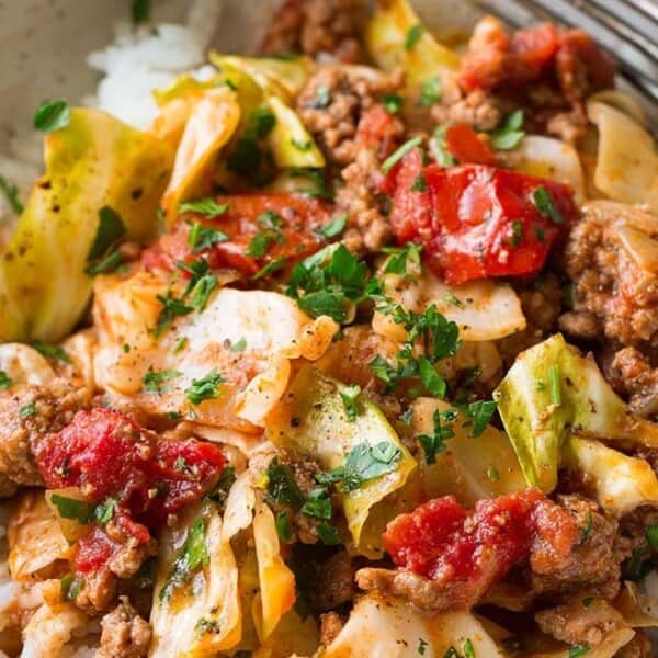 A bowl of rice topped with a colorful mixture of ground meat, cabbage, tomatoes, and herbs. The dish is garnished with fresh parsley and served with a fork on the side.