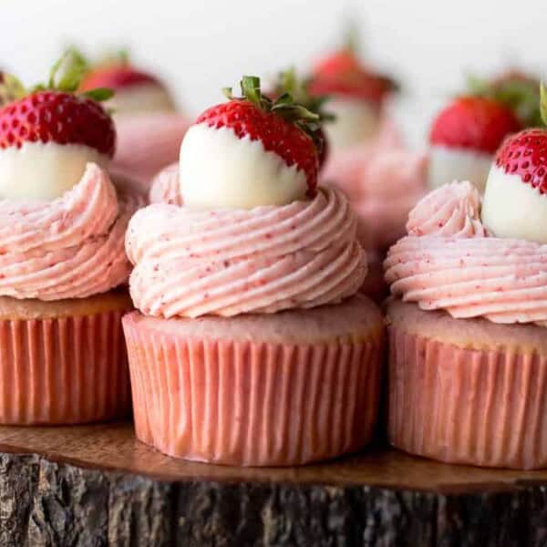 Cupcakes topped with pink frosting and white chocolate-dipped strawberries sit on a wooden slab. The cupcakes have pink wrappers and are arranged in rows, highlighting their decorative toppings and texture.