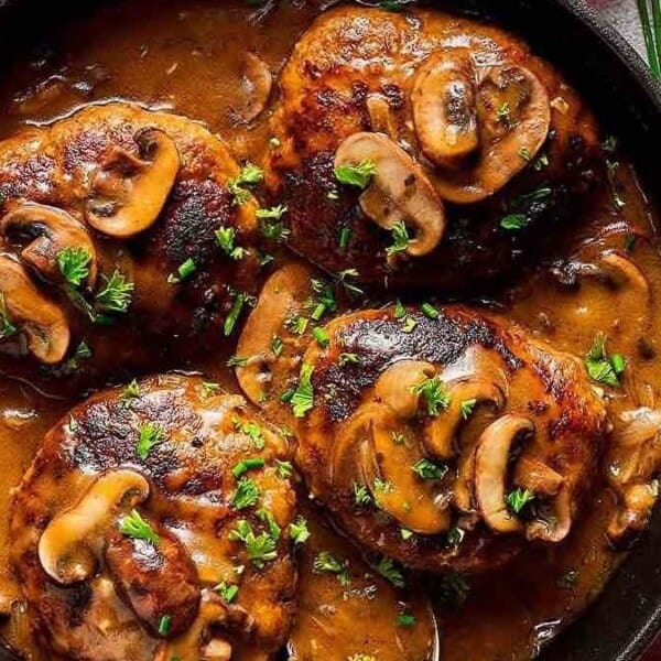 A black skillet filled with four Salisbury steaks topped with sliced mushrooms and garnished with chopped parsley. The steaks are covered in a rich, brown gravy. The skillet is placed on a light surface with a textured towel beside it.
