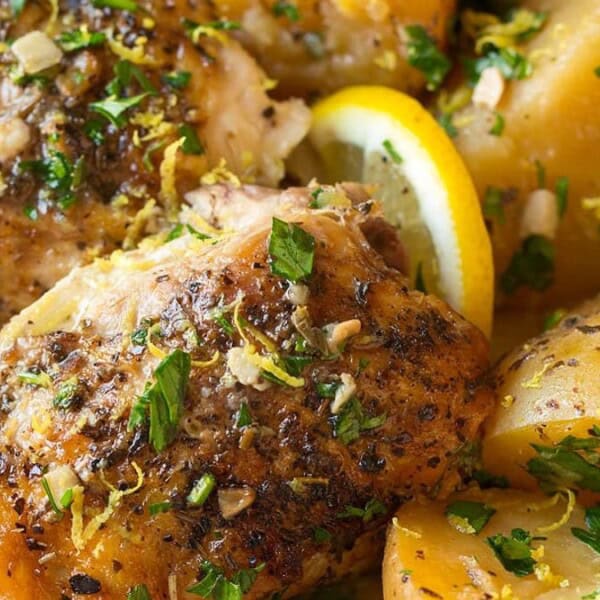 A plate of seasoned roast chicken thighs garnished with herbs and lemon zest, accompanied by golden potatoes and asparagus. A lemon slice adds a touch of freshness to the visually appealing dish.