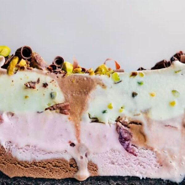 A slice of multi-layered ice cream cake featuring chocolate, vanilla, and strawberry layers, topped with chocolate curls and chopped pistachios, served on a white plate.