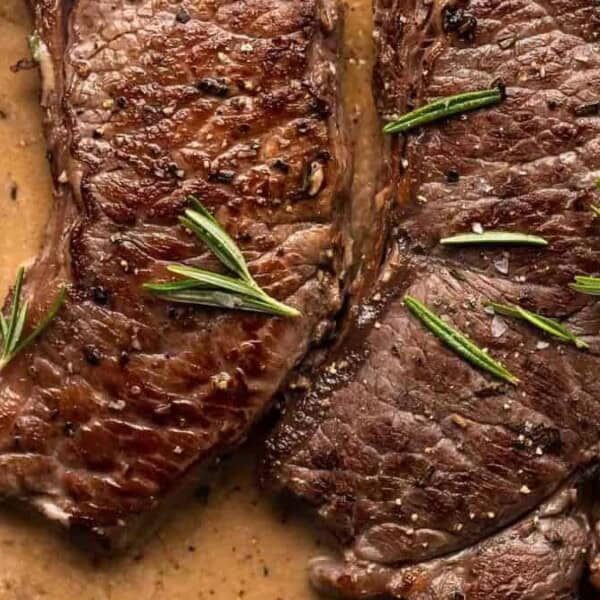 Three seared steaks in creamy sauce, topped with fresh rosemary sprigs, sit in a black skillet.