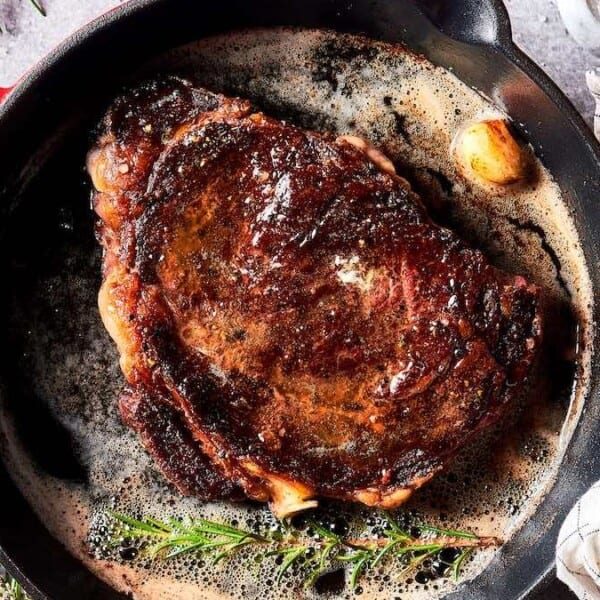 A cooked beef steak in a cast iron skillet with rosemary, garlic, butter, seasonings, and a checkered towel nearby.