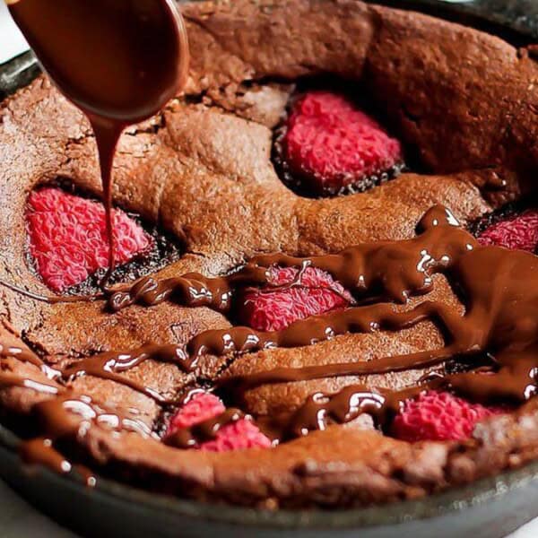A skillet brownie topped with raspberries is drizzled with melted chocolate.