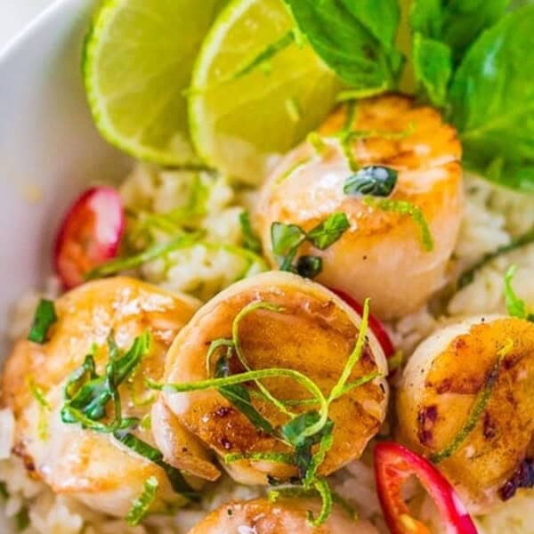 A bowl of rice topped with grilled scallops, sliced red chili, fresh basil leaves, and lime wedges.
