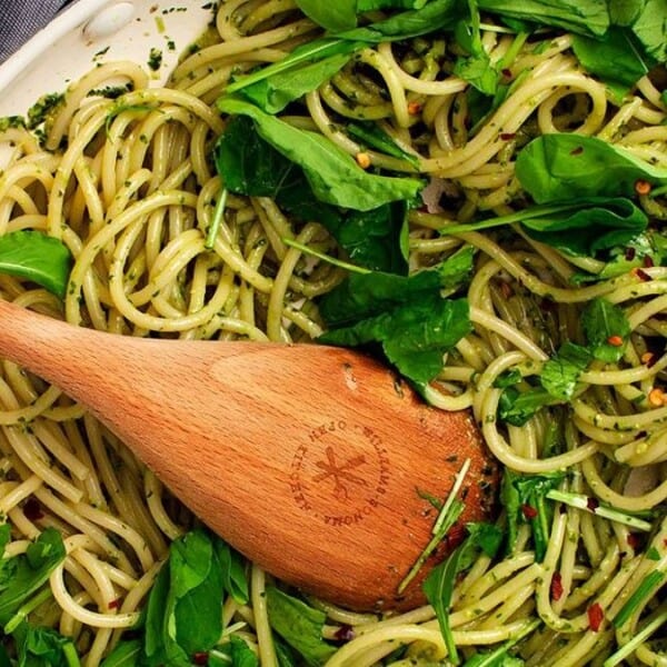 Cooked spaghetti tossed with green pesto and leafy greens in a white pan, being stirred with a wooden spoon.